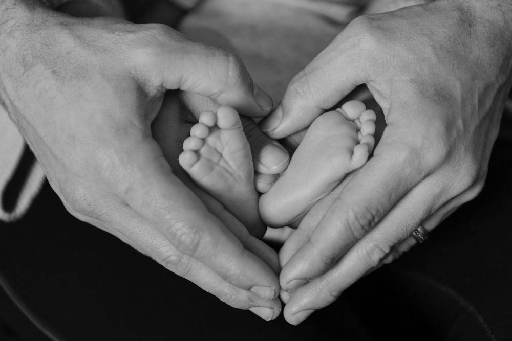 image was here of a father's hands holding the feet of a baby. Hands are shapped like a heart.