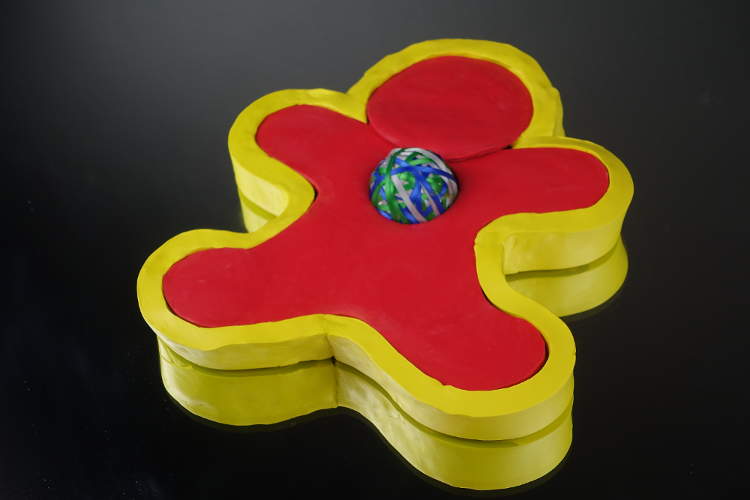 An image was here of a gingerbread man shaped figure in modeling clay. There is a three colored ribbon ball located in the heart region of the gingerbread man. The color surrounding the three color ball is red. Surrounding the red gingerbread man is a yellow layer outlining the entire man.